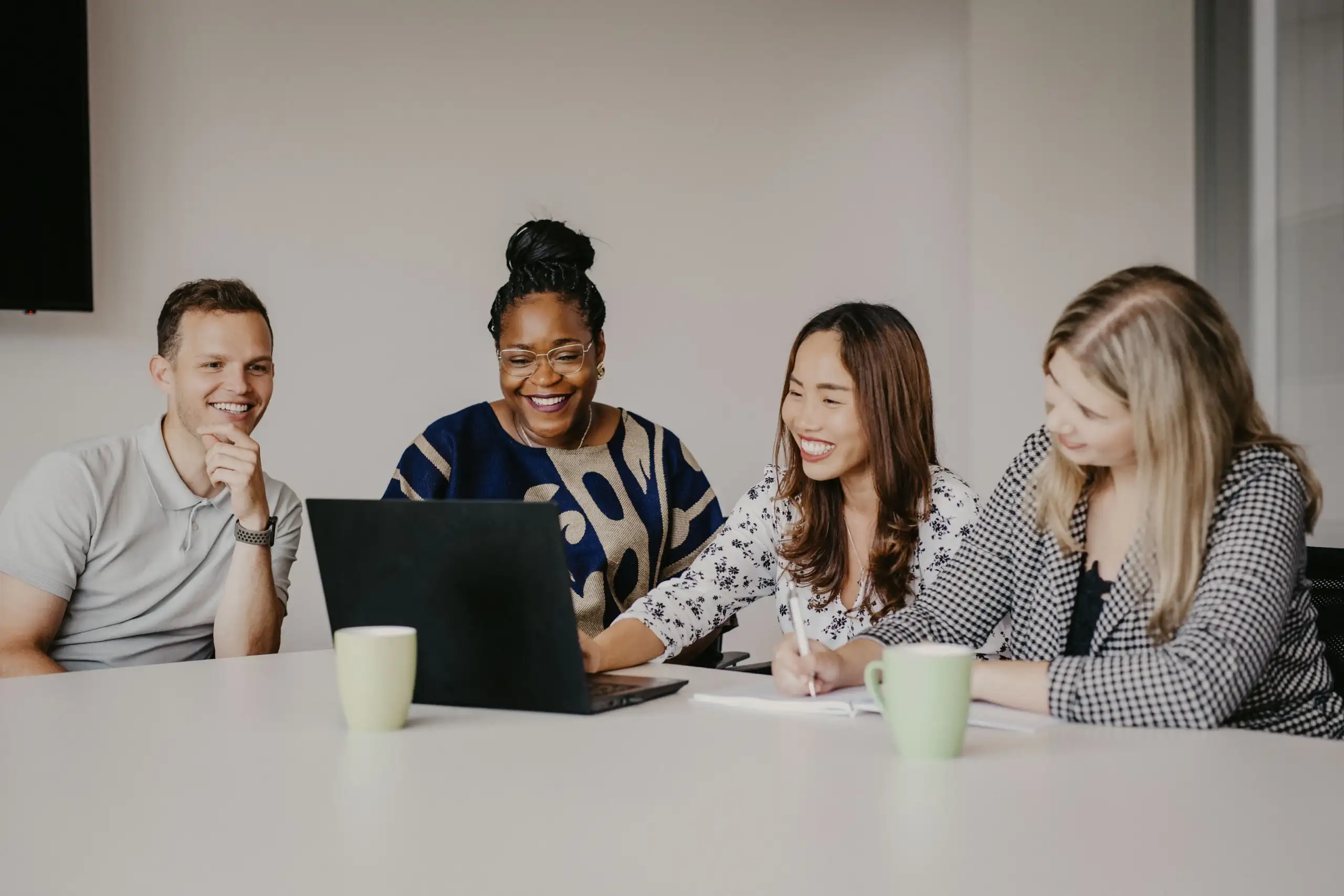 Vier Teammitglieder sitzen gemeinsam an einem Tisch, schauen auf einen Laptop und lachen, während sie an einem Projekt arbeiten.